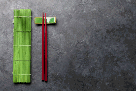 Foto de dois palitos de madeira vermelhos sobre uma esteira verde. Usar hashis para comer é um dos costumes dos japoneses.