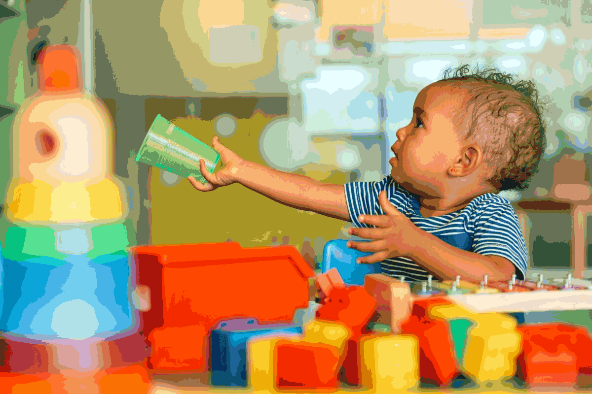 Foto de bebê em meio a brinquedos pedagógicos, segurando copo plástico.  A idade escolar mais adequada para iniciar é uma escolha dos pais, contanto que dentro do estabelecido na lei.