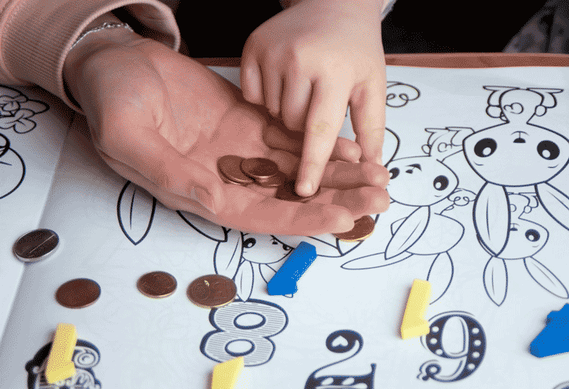 Foto da mão da mãe e do filho com algumas moedas, números e desenhos. O letramento matemático estimula o raciocínio lógico.