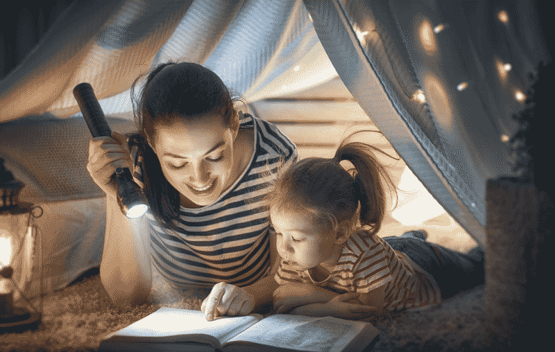 Foto de mãe e filha lendo livro com lanterna em uma barraca dentro de casa. Saber como ensinar a ler começa pelo hábito da leitura.
