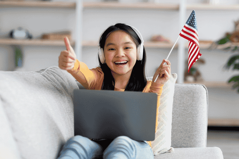 Foto de menina asiática usando fones de ouvindo e fazendo sinal de positivo com as mãos, enquanto sorri e segura uma bandeirinha dos EUA. Saber falsos cognatos é uma grande evolução no aprendizado.