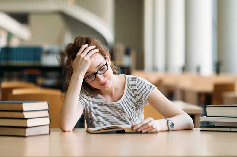 Foto de menina adolescente com a mão na testa, lendo um livro. A dificuldade em matemática é comum entre os alunos.